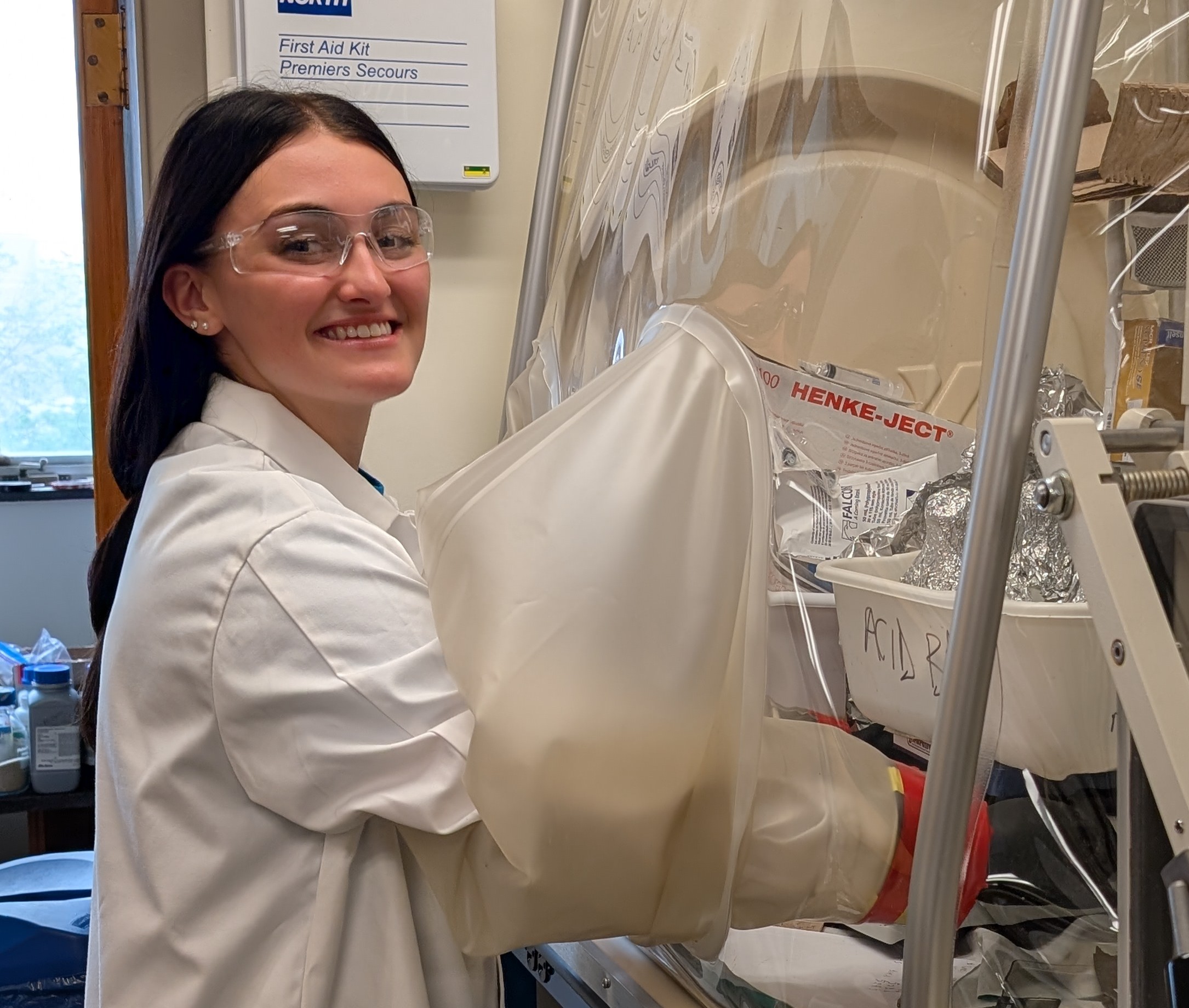 A woman wearing a lab coat and safety goggles standing in front of a glove box