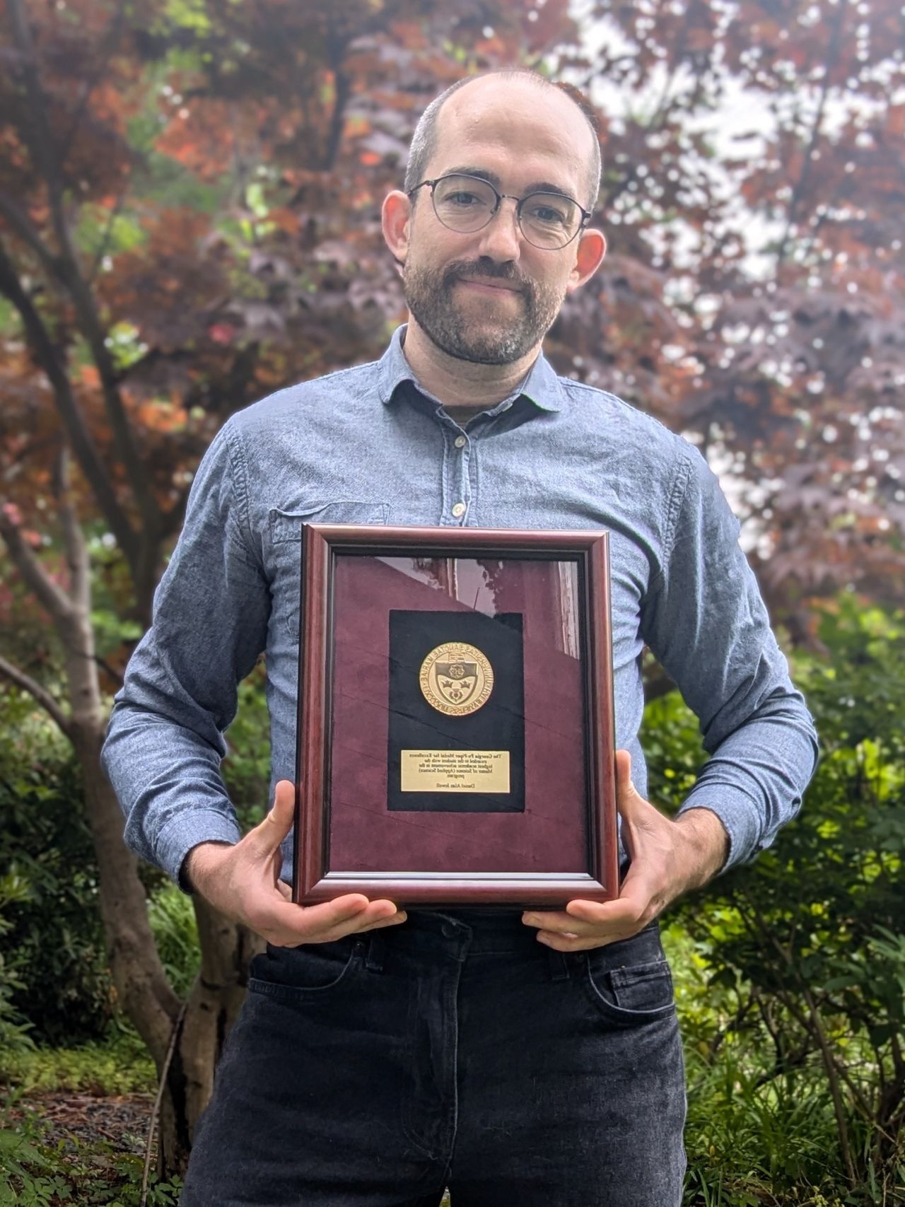 A man is standing in front of some trees outside, holding up the framed Georgia Pe-Piper Medal for Excellence in Applied Science.