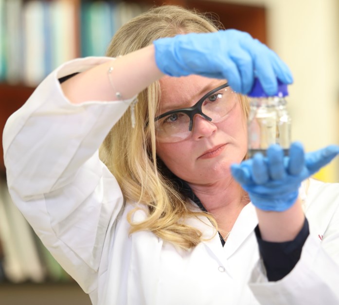 Emily wearing a lab coat and gloves, holding up some glass vials for closer look.