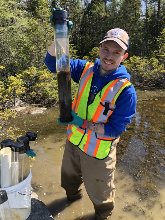 David holding up a sediment column while wearing waders in a wetland