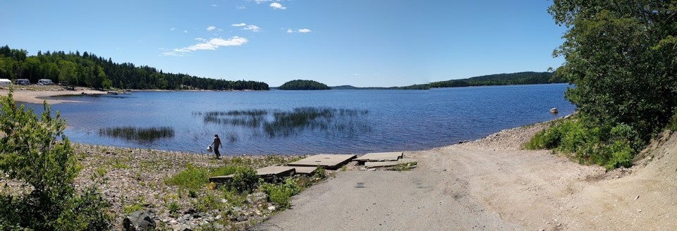 Panorama of a blue lake on a clear day, showing a boat landing road, with a small figure walking along the shoreline