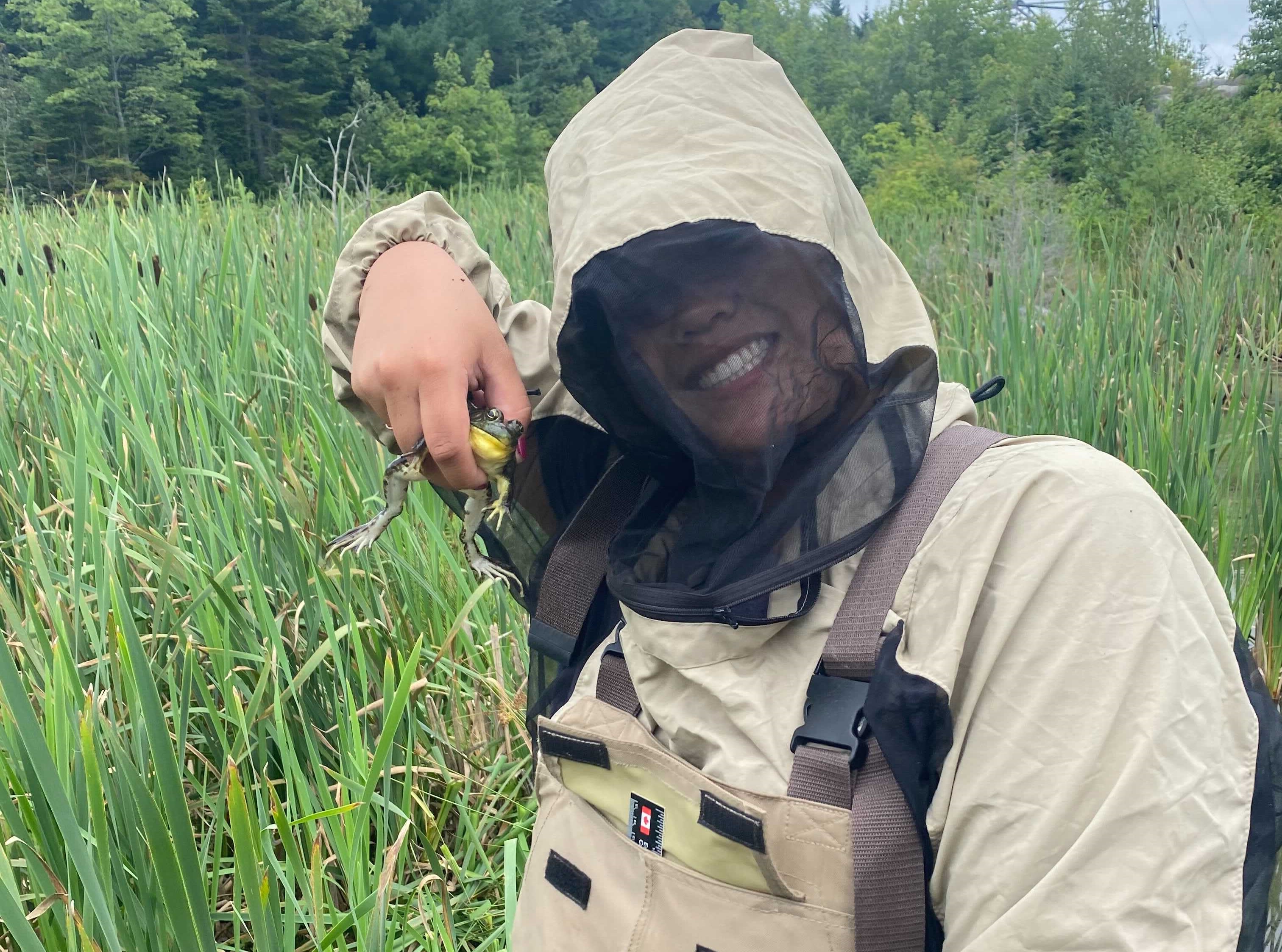 A woman wearing a bug jacket with mesh covering face standing in a green wetland holding up a little frog.