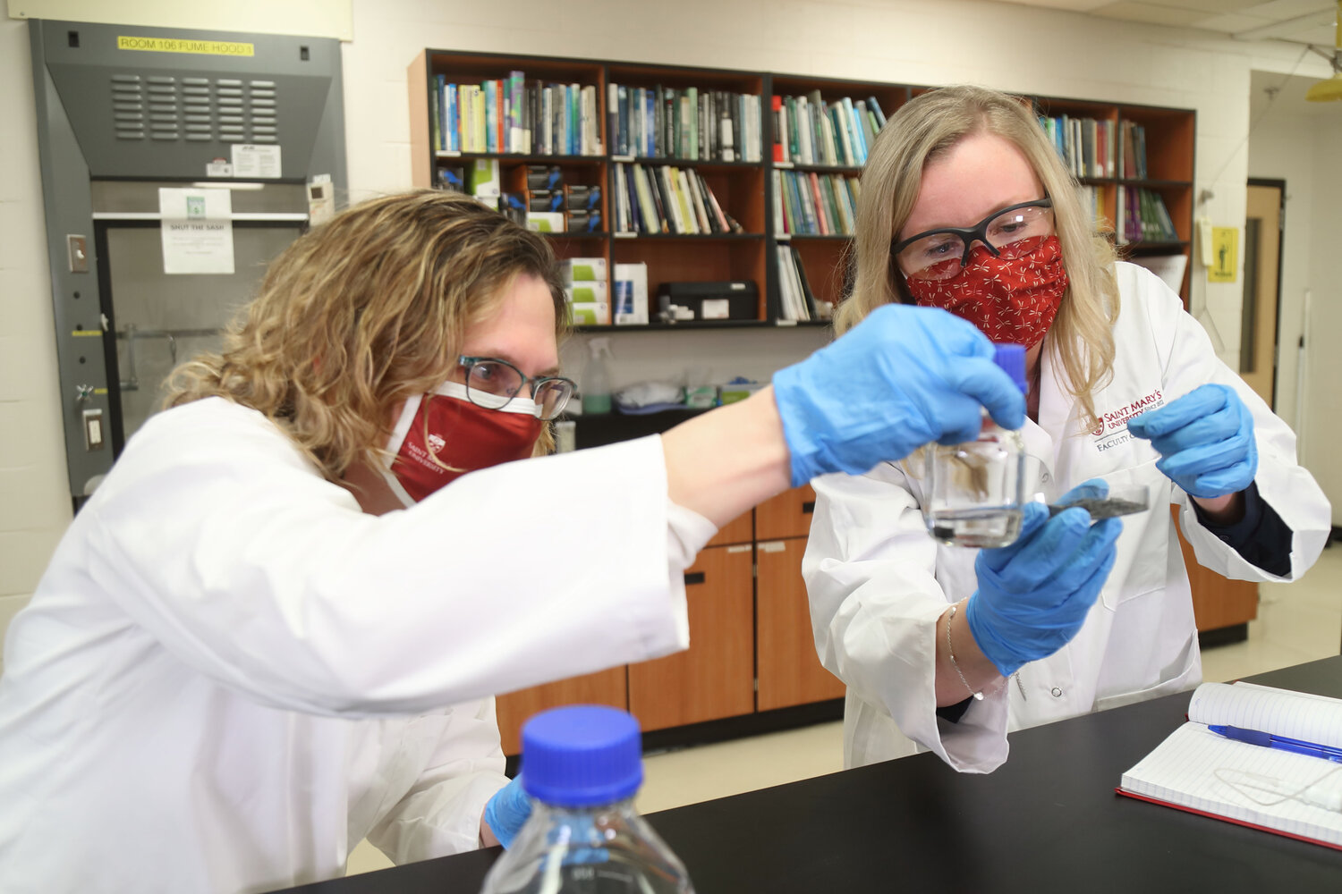 Two light-skinned women wearing red masks and white SMU-logo lab coats posting with some glassware in a laboratory