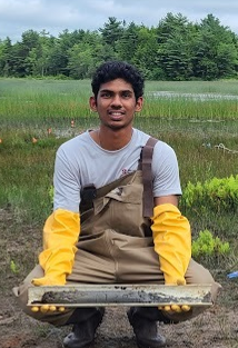 A young man wearing waders and long yellow gloves holding a sediment core