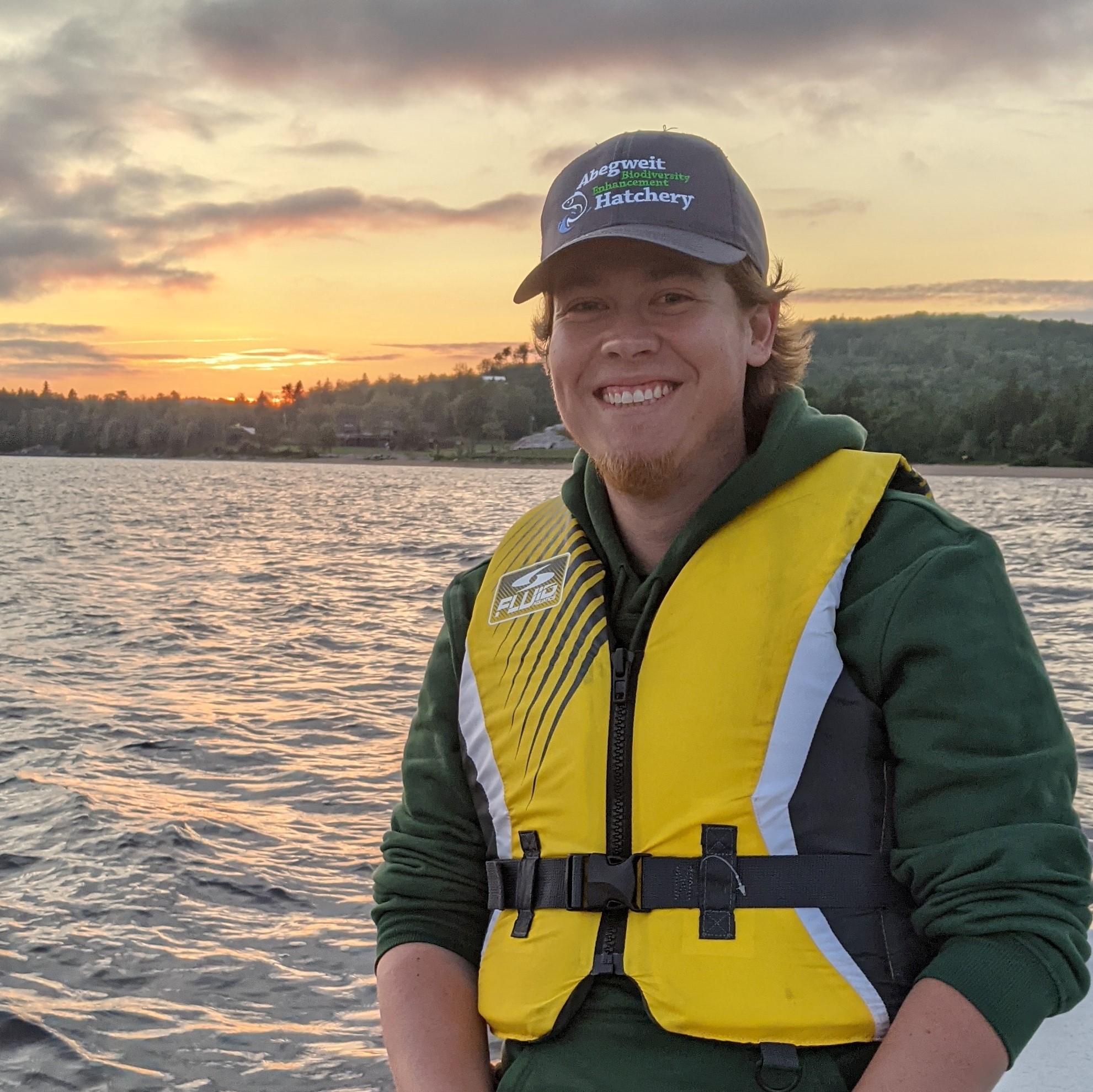 A man sitting on a edge of a small boat wearing lifejacket and ball cap, sunset on a lake behind him