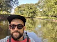 Dan Jewell wearing dark sunglasses in front of a calm waterbody surrouded by leafy trees