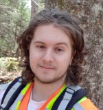 A long-haired person in front of a lichen covered tree trunk, wearing safety vest and harness