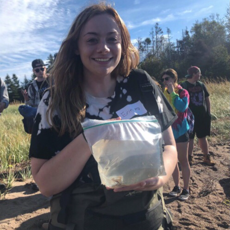 A lady with long brown hair wearing field clothing holding a ziploc bag with water sample