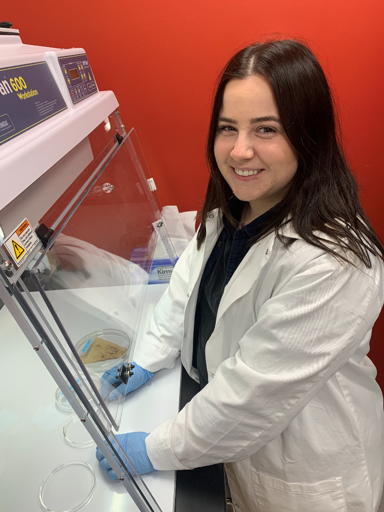 Heidi at Work in front of biosafety cabinet in white lab coat, with bright red background