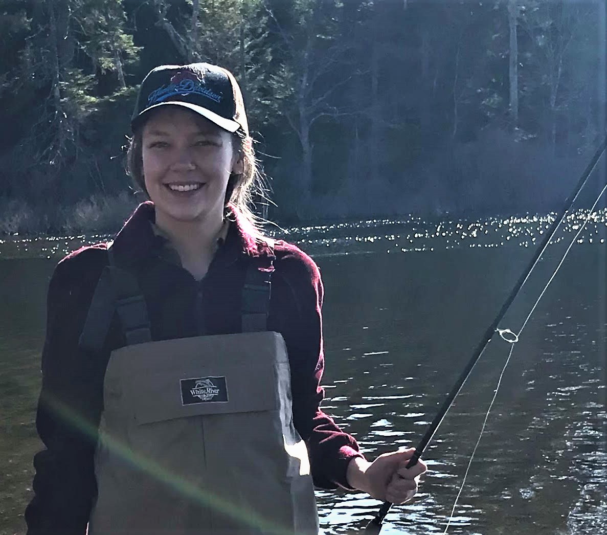 Jenna wearing waders and fleece jacket in a river, holding a fishing rod