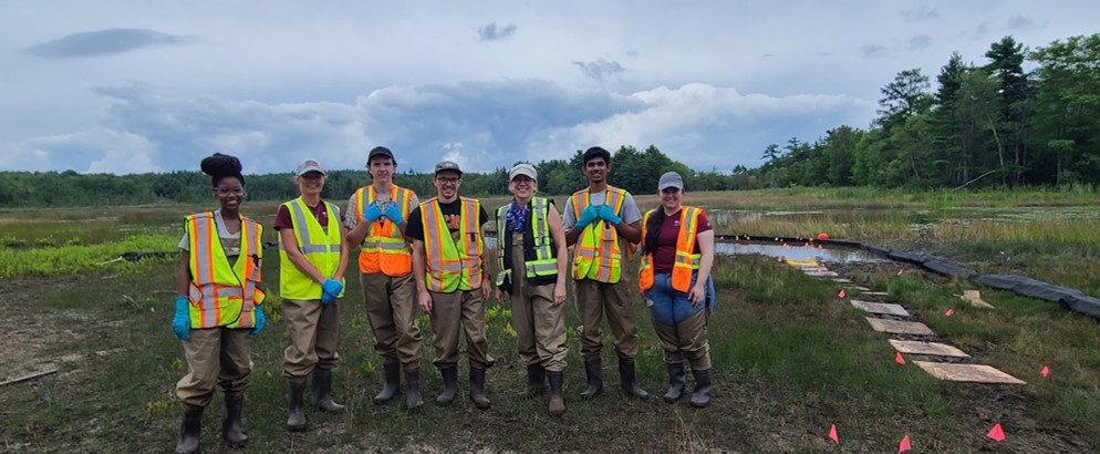 A group of tired and smiling people wearing waders and safety vests in front of a muddy wetland