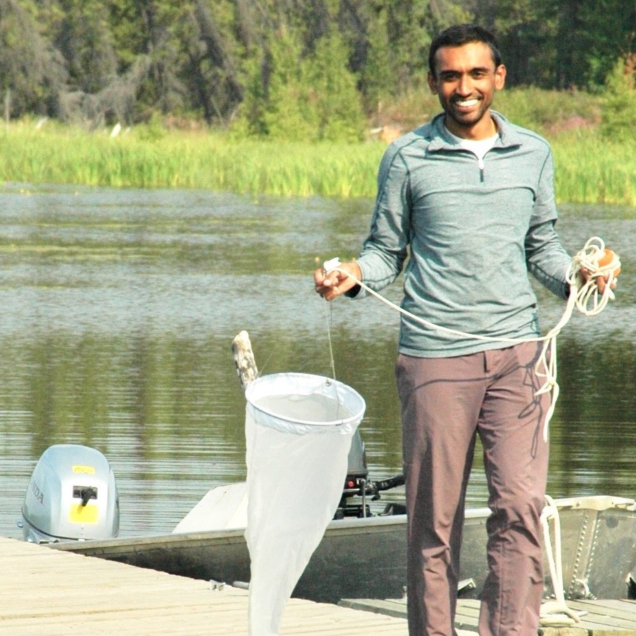 A man wearing field clothing holding a zooplankton net standing on a dock in a boreal lake